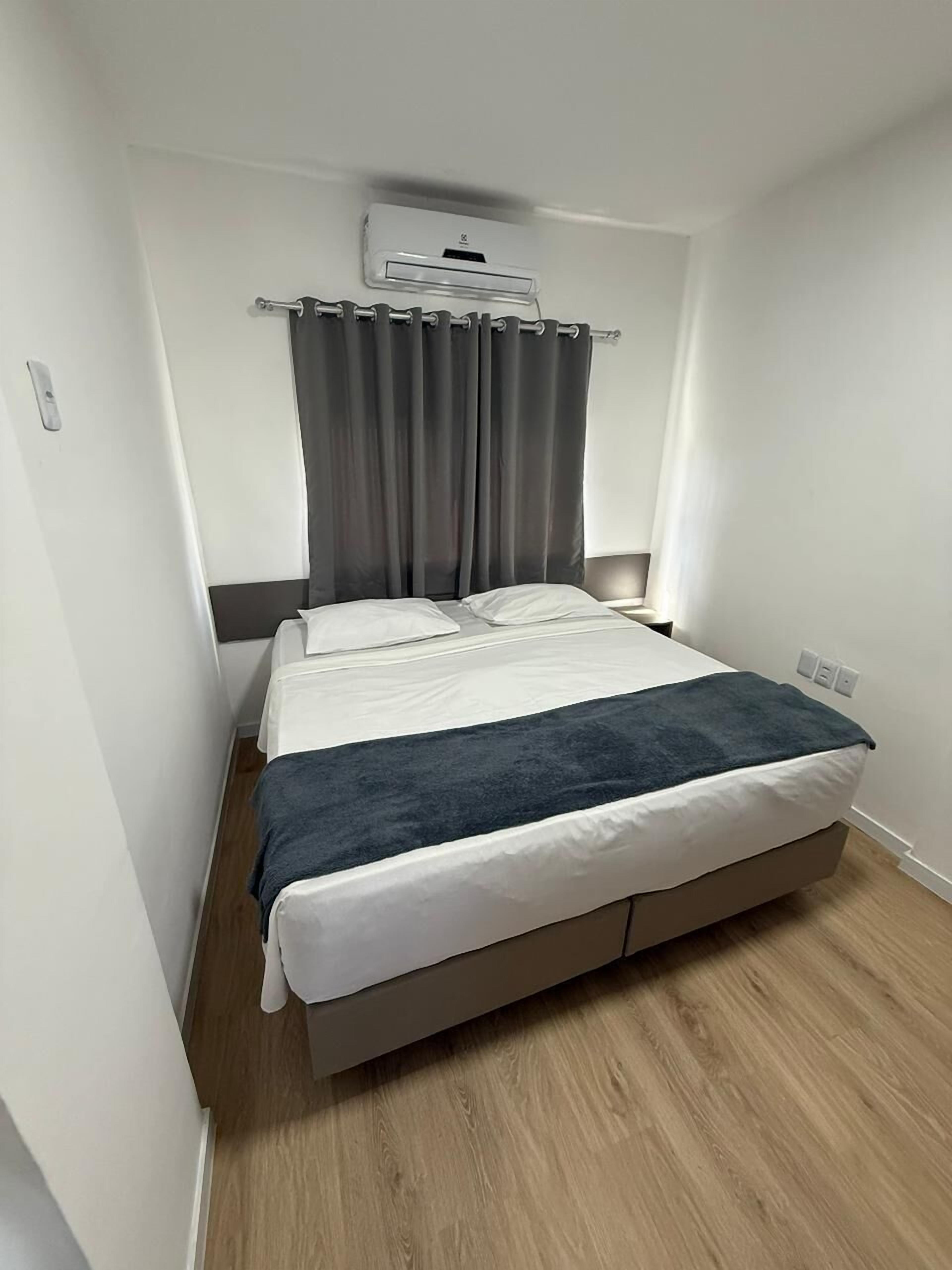 Una camera singola con un letto e una testiera grigia, una coperta grigio e bianca, una coperta grigia, un pavimento in legno e una finestra con tende grigie.