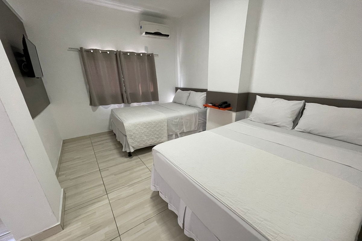 A hotel room with a large bed, two smaller beds, a TV, an air conditioner, and a curtain.