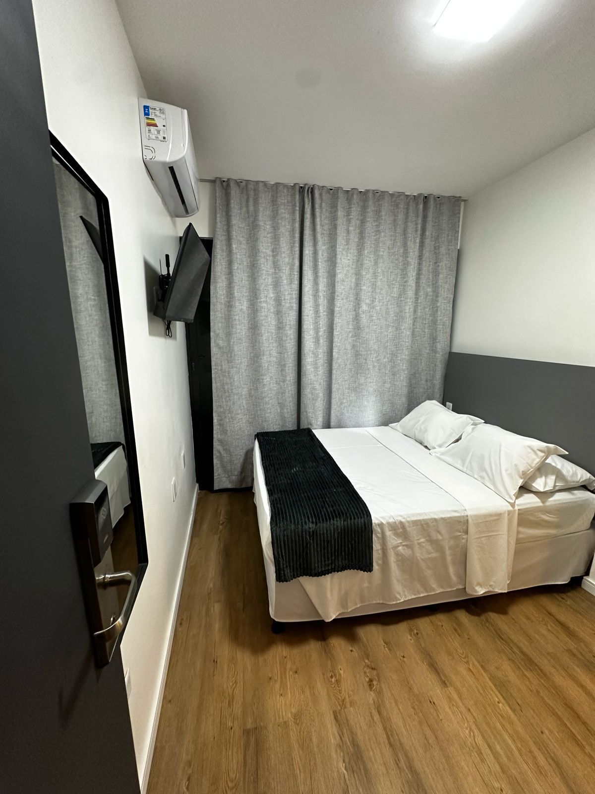 Quarto com cama, ar-condicionado fixado na parede e janela com cortinas cinzentas.