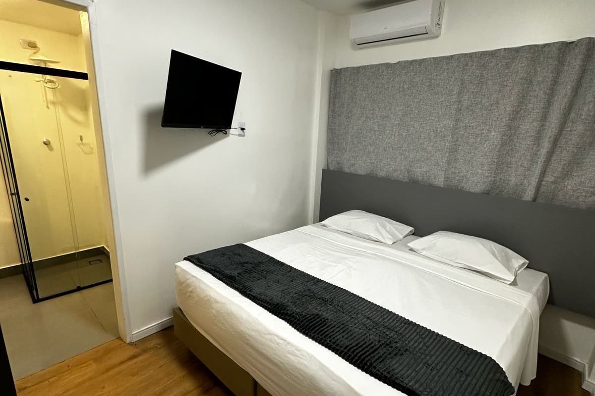 A hotel room with a bed, a television mounted on the wall, an air conditioning unit, and a door leading to a bathroom.