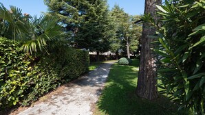 A paved pathway flanked by lush greenery and a large tree. - Residence Sant Elena A19 by Wonderful Italy (Peschiera del Garda)