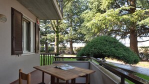 A balcony with a wooden table and chair, a small garden with a bush, and a view of trees and a body of water. - Residence Sant Elena A19 by Wonderful Italy (Peschiera del Garda)