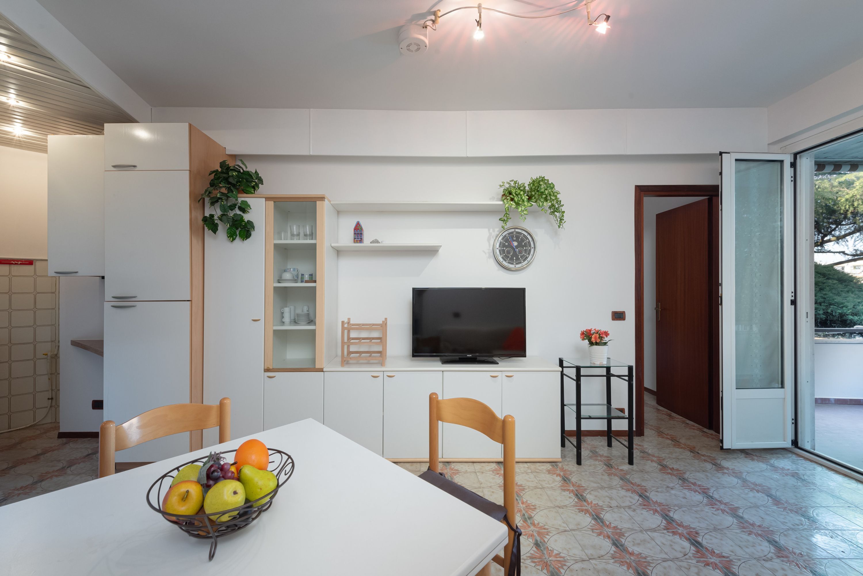 Una cucina moderna con mobili bianchi, un tavolo da pranzo con sedie, una ciotola di frutta, un televisore e una piccola pianta.