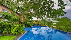 Una piscina con borde de azulejos, rodeada de exuberante vegetación y un edificio al fondo.