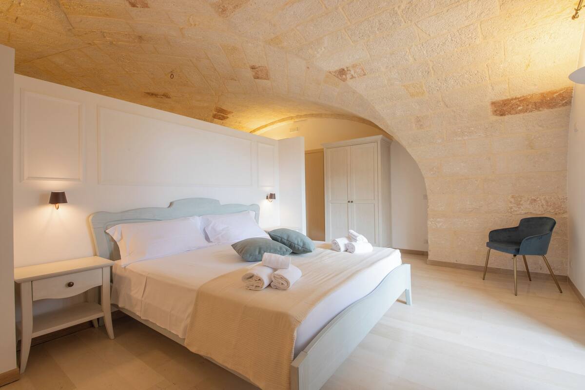 Ein Schlafzimmer mit Steindecke, einem Bett mit weißer Bettwäsche, einem Nachttisch, einem Sessel und einem Kleiderschrank.