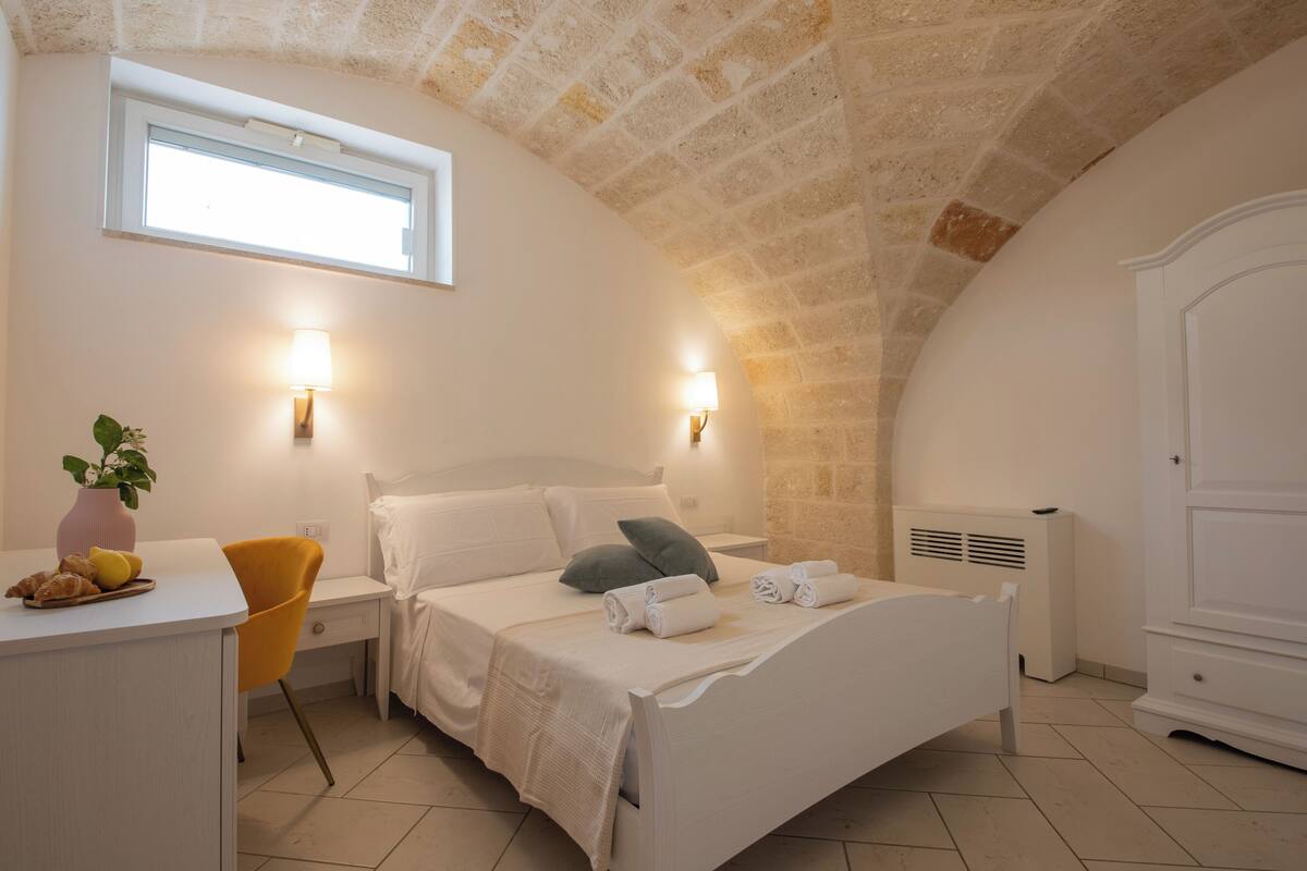 Ein Schlafzimmer mit Bett, Nachttischlampen, Schreibtisch mit Stuhl, Heizkörper und Fenster.