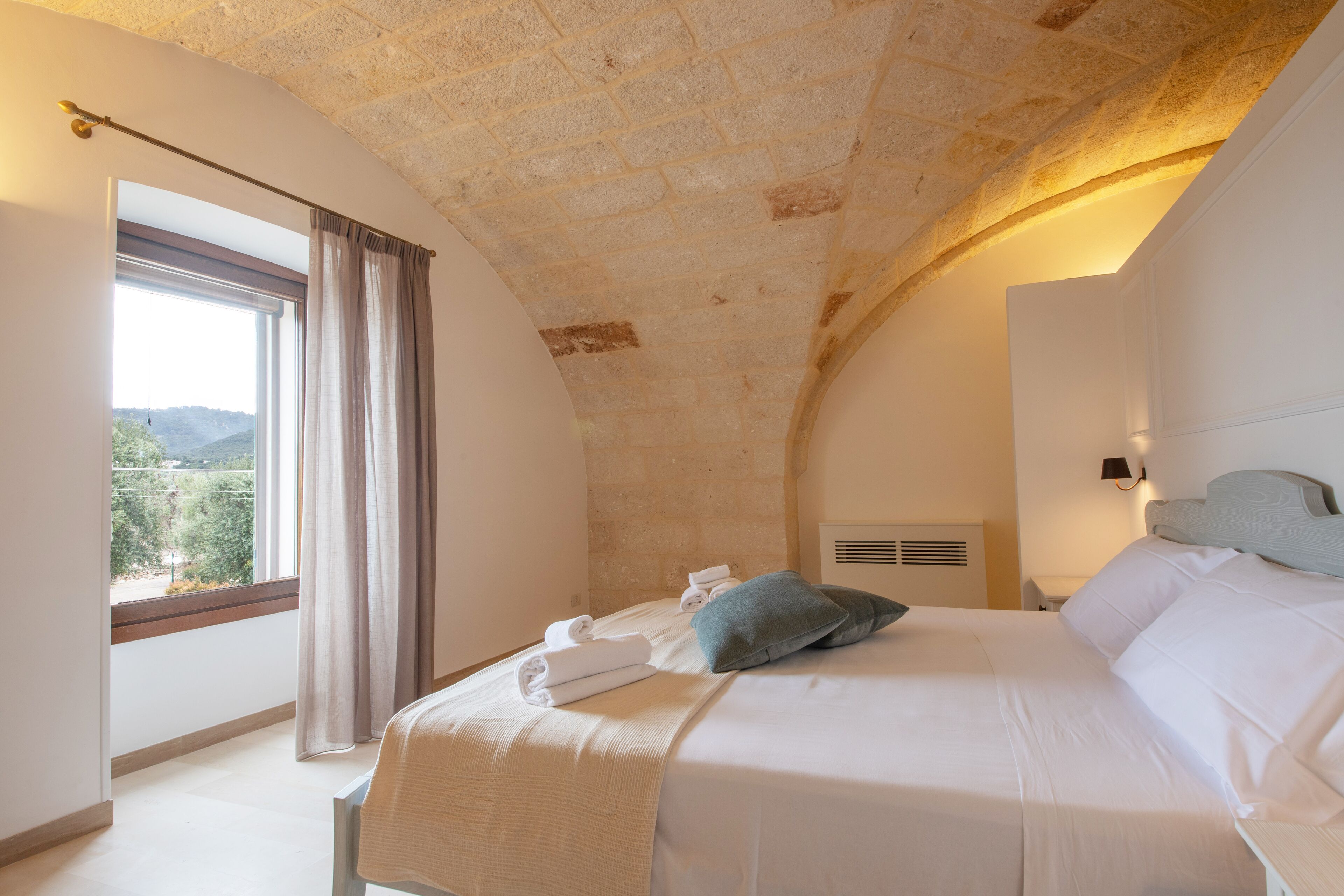 Ein Schlafzimmer mit Steindecke, einem großen Bett mit weißer Bettwäsche, einem Ausblick ins Freie durch ein Fenster und einem Nachttisch mit Lampe.