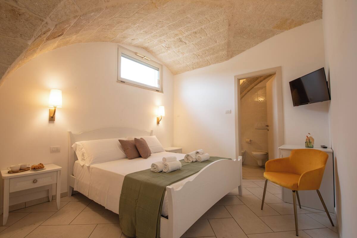 Ein Schlafzimmer mit Bett, Nachttischen, einem Sessel, einem Fernseher und einem durch einen Bogen sichtbaren Badezimmer.