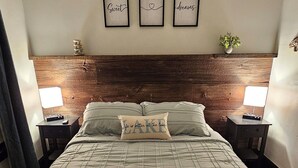 Un lit bien fait, recouvert d’une courtepointe matelassée, deux tables de chevet avec des lampes, une tête de lit ornée d’un panneau en bois et trois citations inspirantes encadrées accrochées au mur.