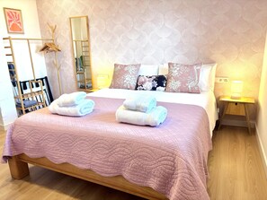 Een keurig opgemaakt bed met een roze deken, twee stapels handdoeken, een houten nachtkastje met een lamp en een spiegel aan de muur.