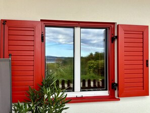 Une fenêtre avec des volets rouges, une vue sur la verdure et des bâtiments éloignés, et un ciel dégagé.
