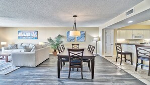 A spacious living room with a dining area, a kitchen, and a door leading to another room.
