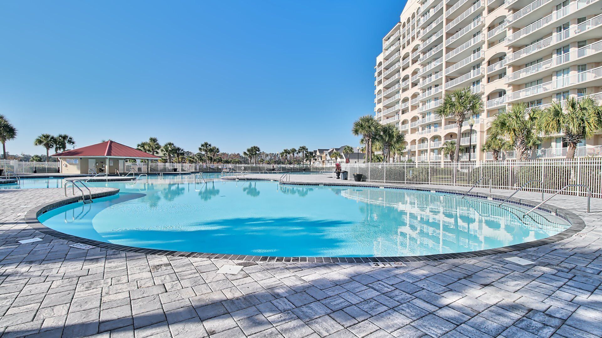Une grande piscine extérieure entourée d’une promenade dallée, avec un immeuble d’hôtel à plusieurs étages en arrière-plan.