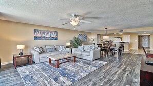 A spacious living room with a sofa set, a coffee table, and a dining area with chairs and a table. A ceiling fan and a painting of sailboats are visible.