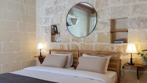 Un dormitorio con pared de piedra, un espejo redondo, mesitas de noche con lámparas y una cama con ropa de cama blanca.
