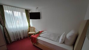 A hotel room with a single bed, a chair, a TV mounted on the wall, and a small table with a remote control. - Room in Guest Room - Comfortable Single Room With Shared Bathroom and Kitchen (Forbach)