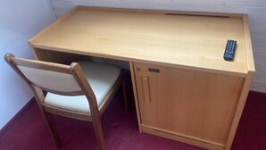 Un petit bureau en bois, une chaise, une télécommande et un meuble de rangement.