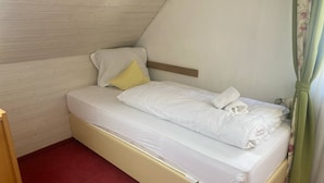 Une petite chambre avec un lit simple, un plafond en pente, une tête de lit en bois, une moquette rouge et un rideau vert.