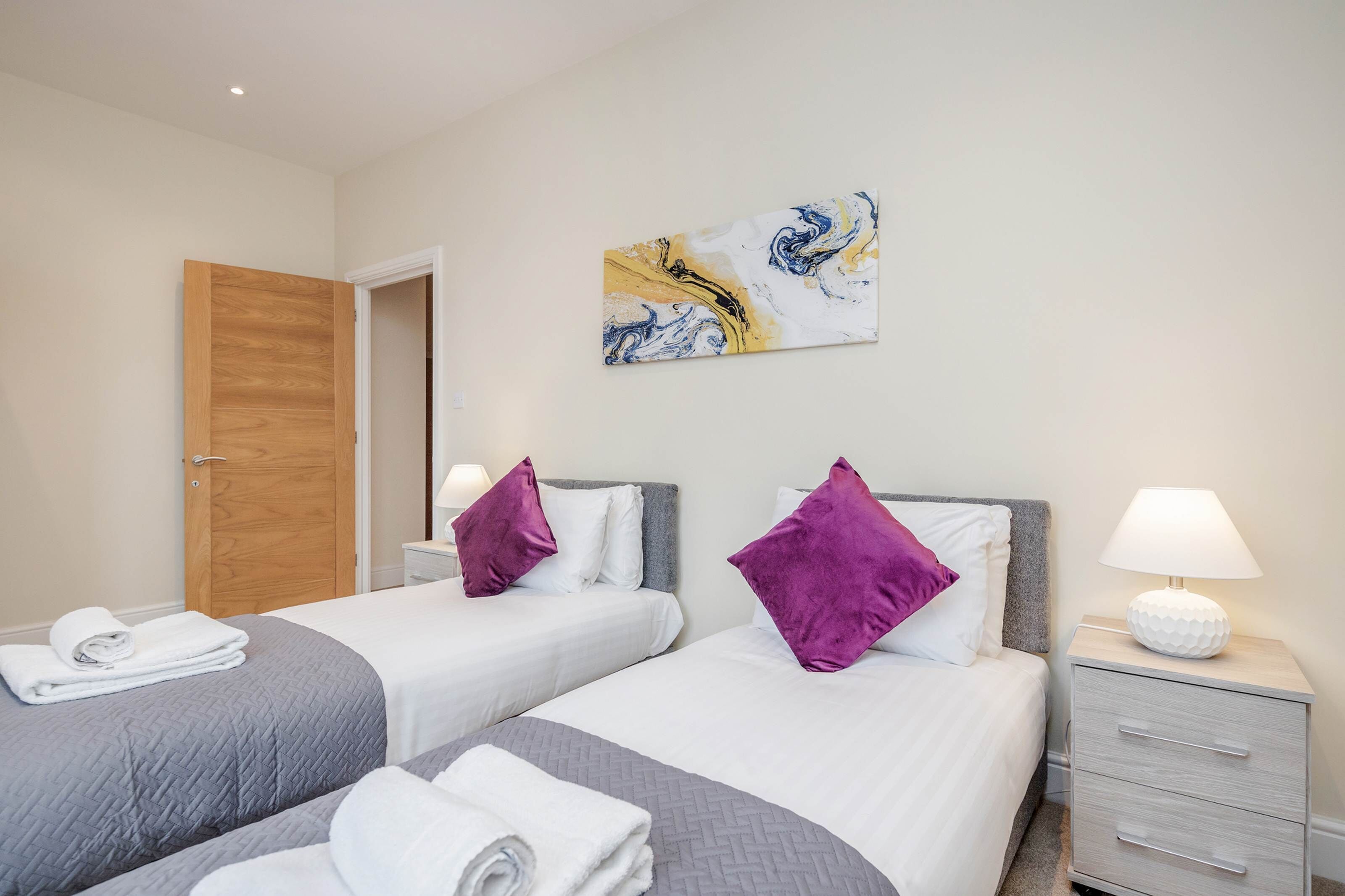 Een hotelkamer met twee bedden, een houten deur, een schilderij aan de muur en een bedlampje.