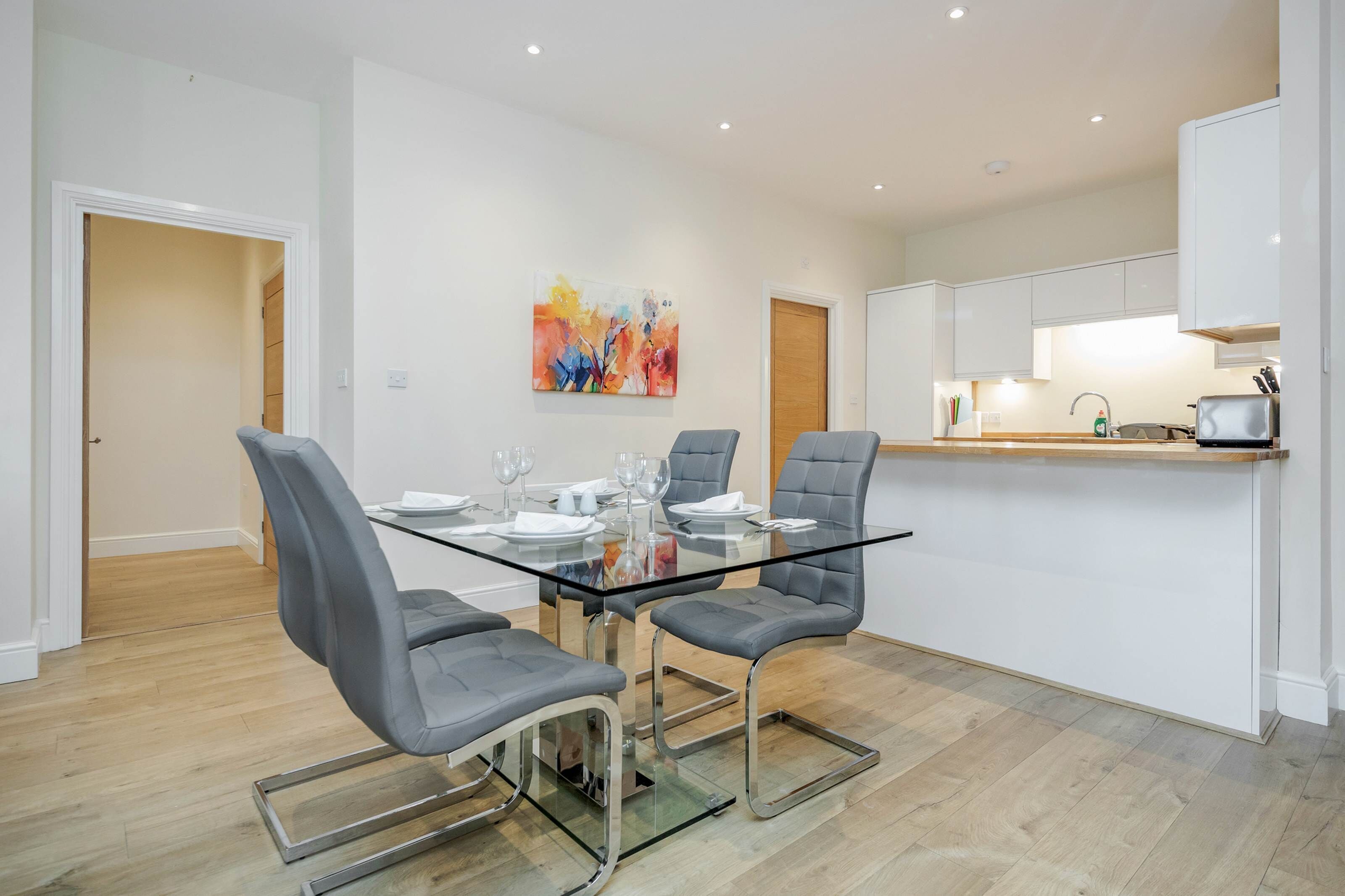 A modern kitchen with white cabinets, a glass dining table, and grey chairs.