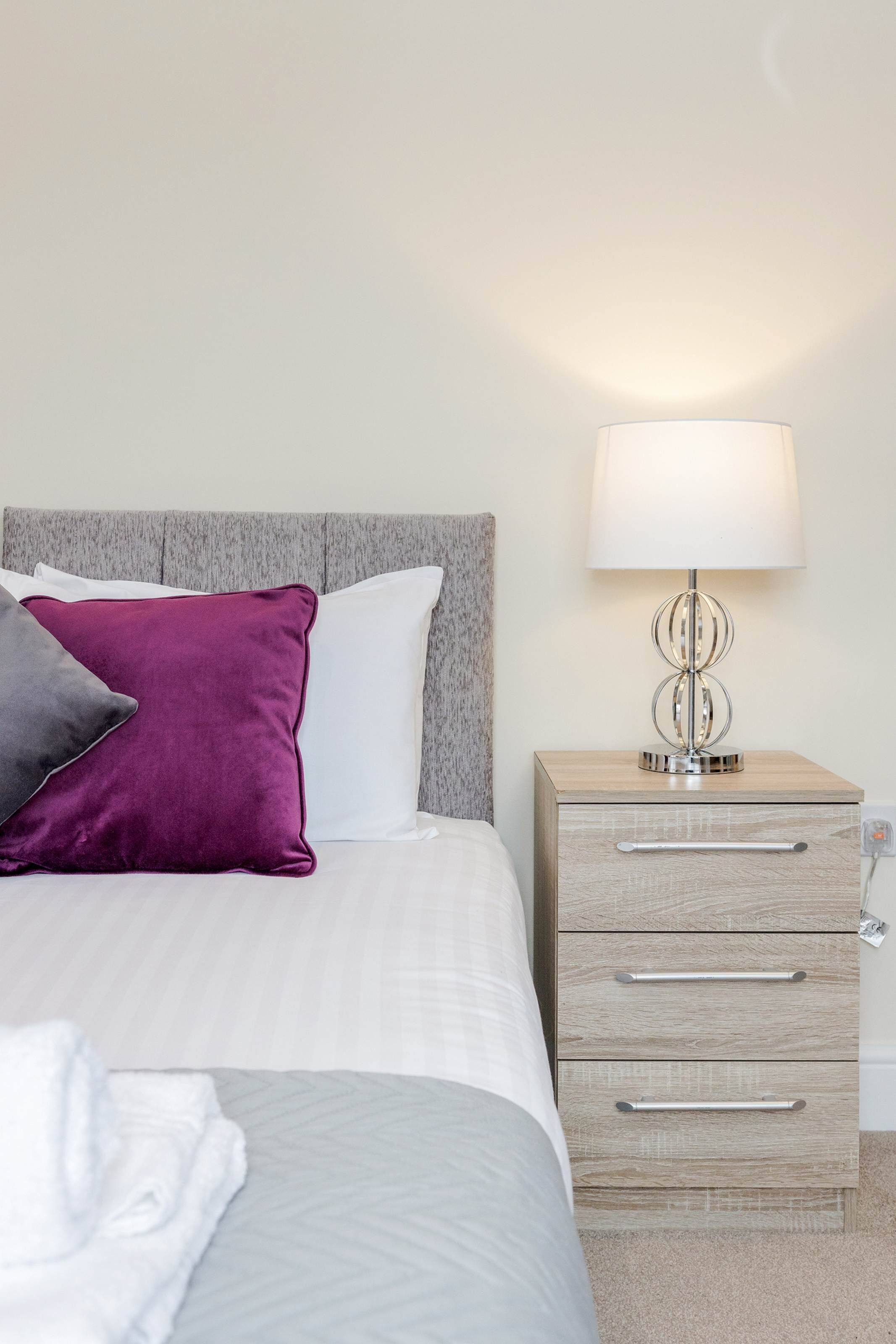A neatly made bed with white and gray bedding, a purple and gray pillow, and a bedside table with a lamp.