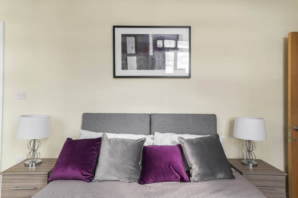 A neatly made bed with a grey headboard, flanked by two bedside tables with lamps, and a framed abstract artwork above.
