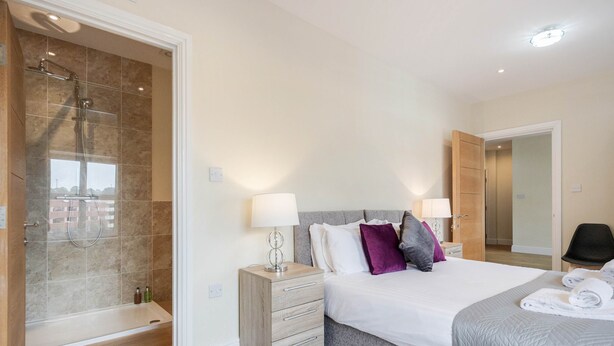 Una habitación de hotel moderna con ducha, una cama con colcha gris y una mesita de noche de madera.