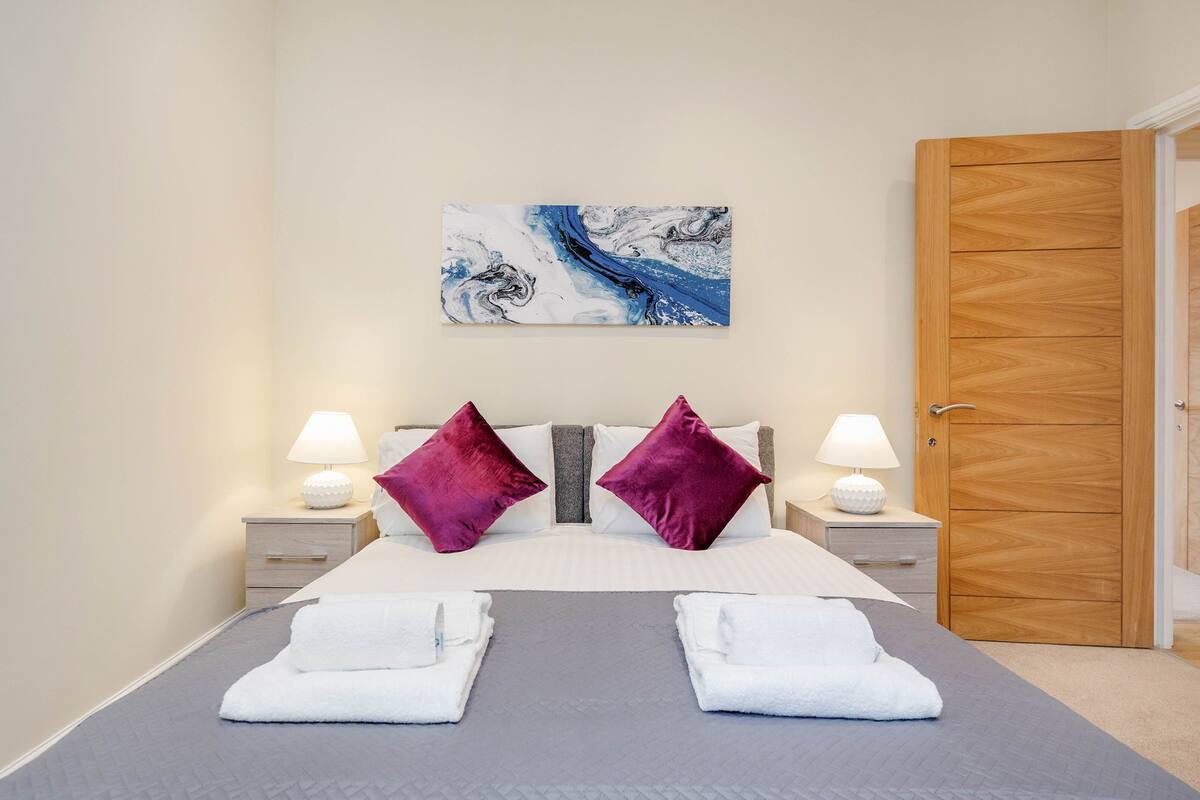 A neatly made bed with two pillows, a bedside table with a lamp, and a wooden door.