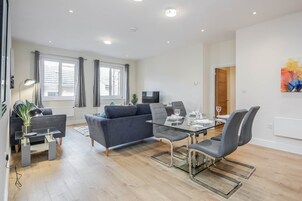 A modern living room with a glass dining table, grey sofas, and a flat-screen TV.