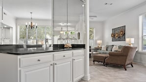 A spacious kitchen with white cabinetry and a black countertop, a dining area with a brown armchair and a table, and a large window with blinds.