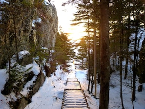 Un sentier en bois recouvert de neige, bordé d’arbres et de falaises rocheuses, qui mène vers un arrière-plan baigné de soleil.