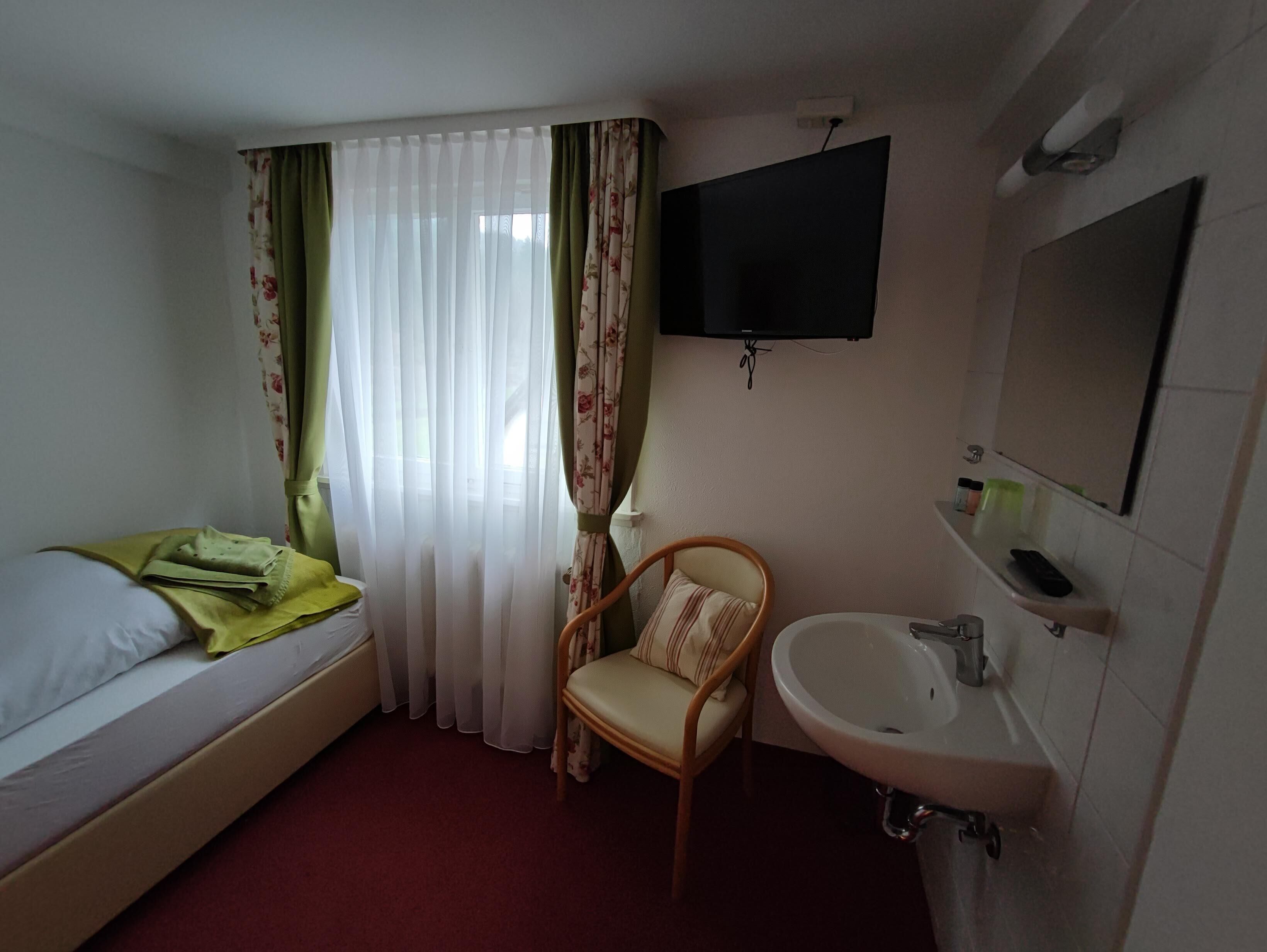 Ein kleines, sauberes Zimmer mit einem Einzelbett, einem Sessel, einem Waschbecken und einem an der Wand befestigten Fernseher.