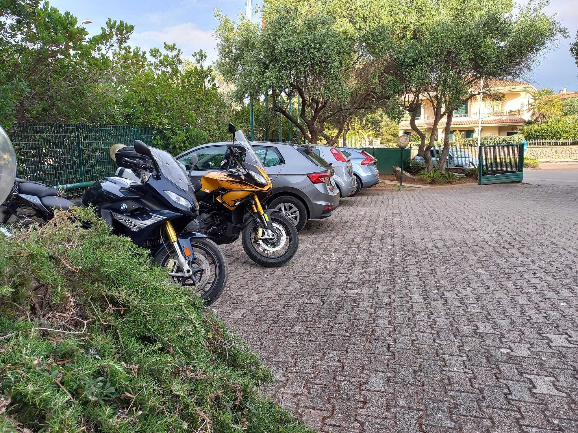 A paved pathway with parked motorcycles and cars, surrounded by greenery and a fence.