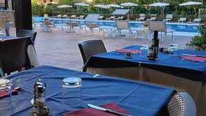 An outdoor dining area with tables set for two, overlooking a swimming pool and a building with balconies. - Green Sporting Club Hotel (Alghero)