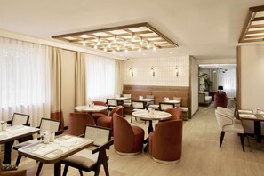 A modern dining area with round tables and cushioned chairs, a wooden ceiling with recessed lighting, and a view into a living area with a sofa and curtains. - The Marmara Çamlıca (Istanbul)