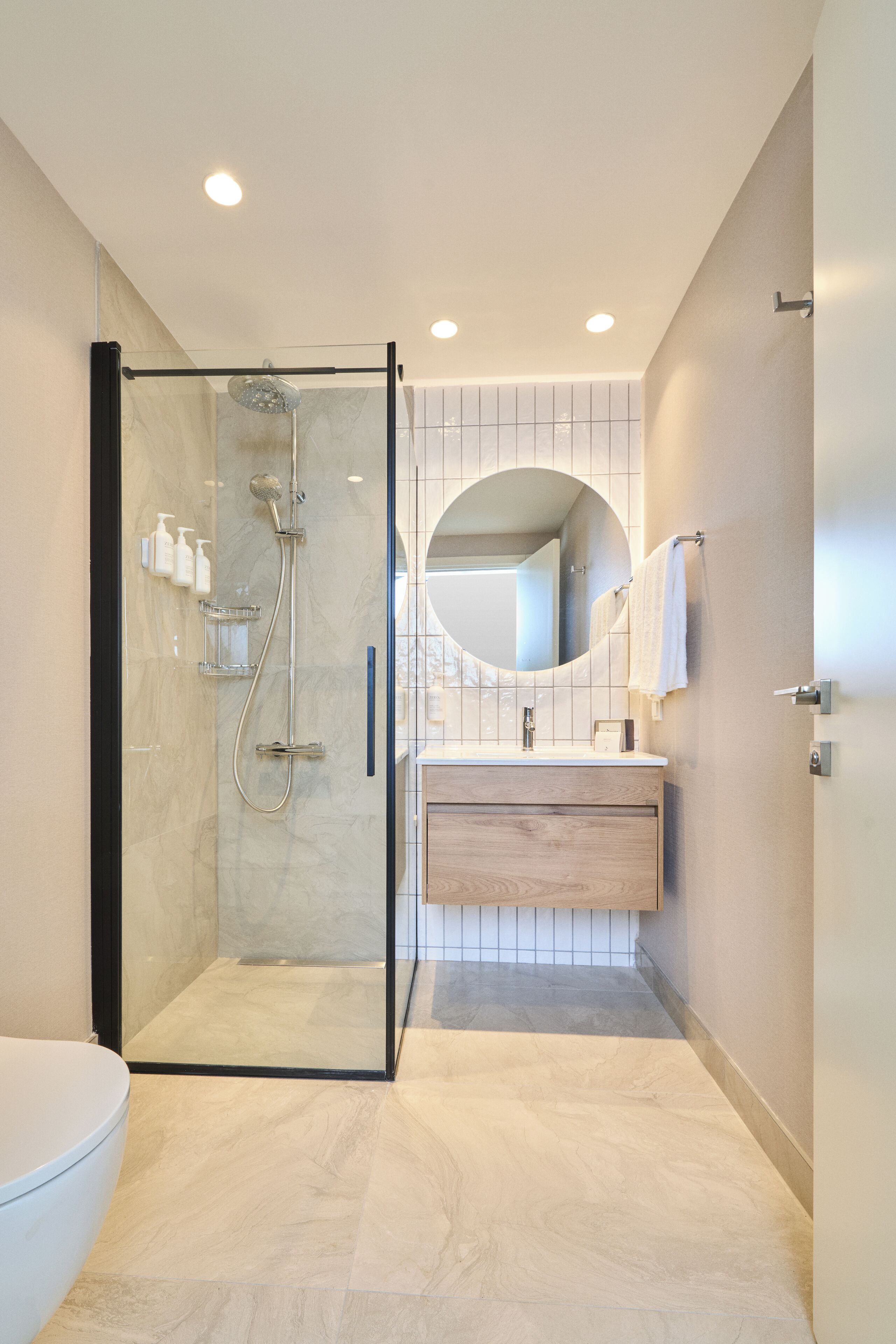 Een moderne badkamer met een douchecabine van glas, een houten wastafelmeubel en een ronde spiegel.