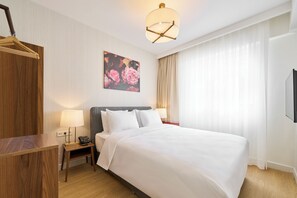 Una habitación de hotel con una cama grande, una mesita de noche con lámpara, un escritorio de madera, un cuadro en la pared y una lámpara del techo.