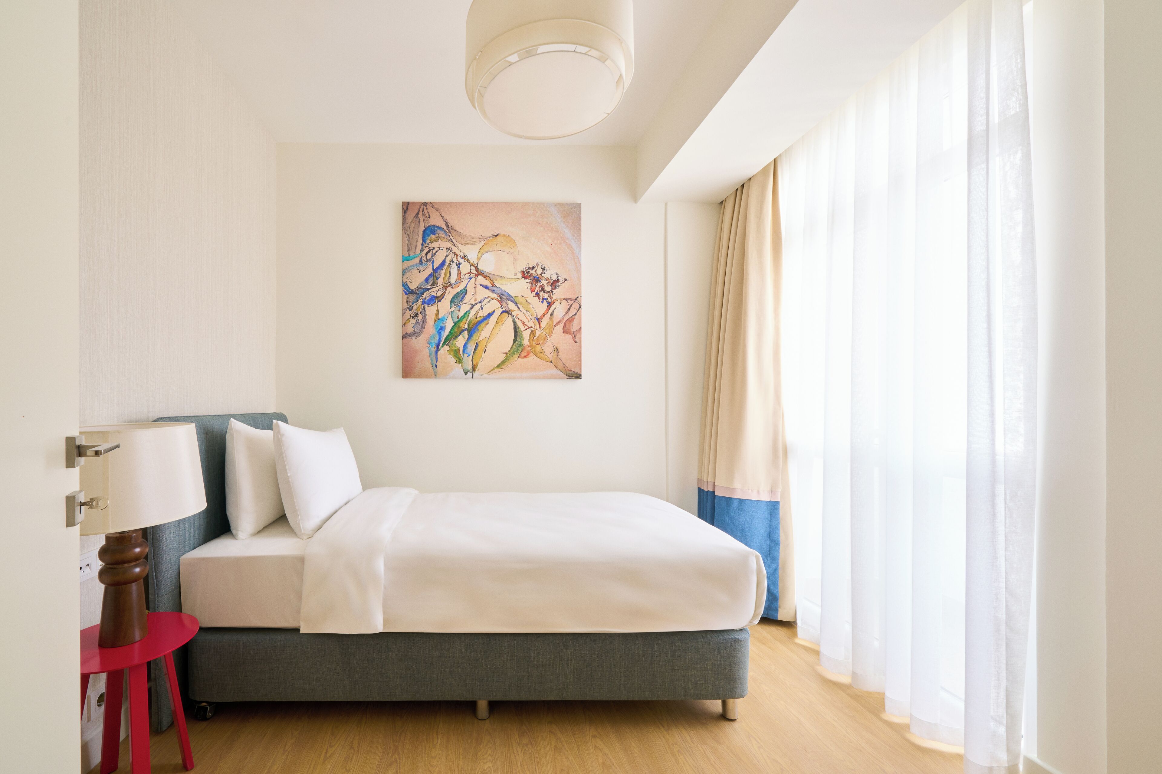 Habitación de hotel ordenada con una cama, una mesita de noche con lámpara, un cuadro en la pared y cortinas traslúcidas en la ventana.