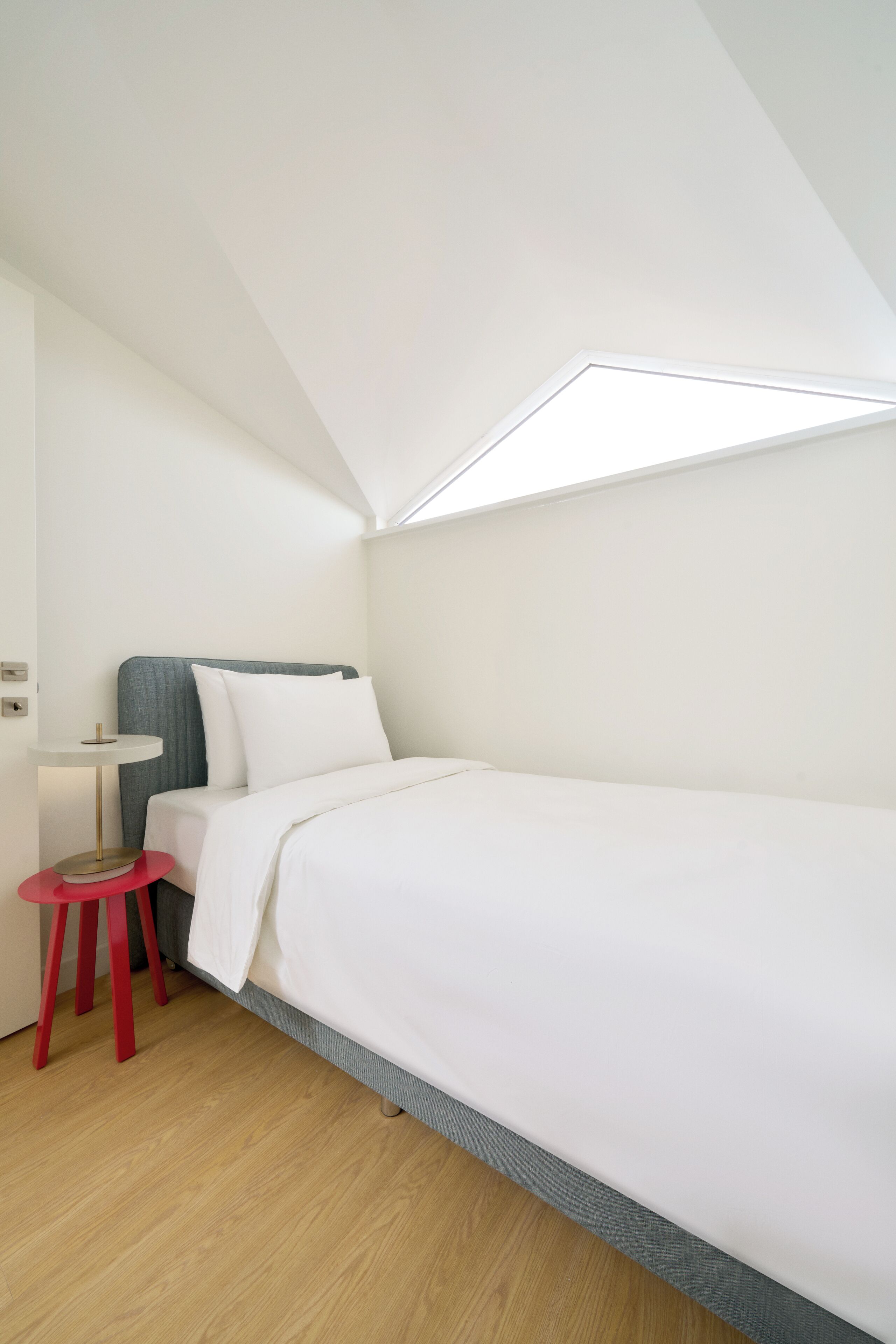 Une chambre compacte et moderne, avec un lit simple, un petit tabouret rouge et une table de chevet blanche.