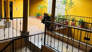 Un couloir aux murs jaunes, doté d’un sol carrelé, de plantes en pot et d’une balustrade de balcon.