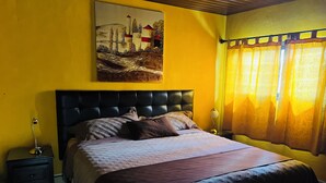 Une chambre à coucher avec une tête de lit noire recouverte de tissu, un lit avec une couvre-lit à motifs, une table de chevet avec une lampe et un tableau accroché au mur.