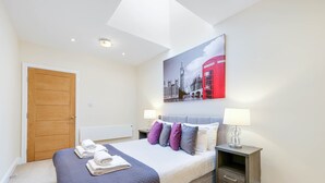 A hotel room with a bed, a nightstand, a lamp, a painting of a red telephone booth, and a wooden door. - Luxury 3 Bedroom Apartment with Parking (Maidenhead)