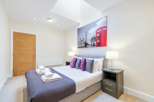 A hotel room with a bed, a nightstand, a lamp, a painting of a red telephone booth, and a wooden door.