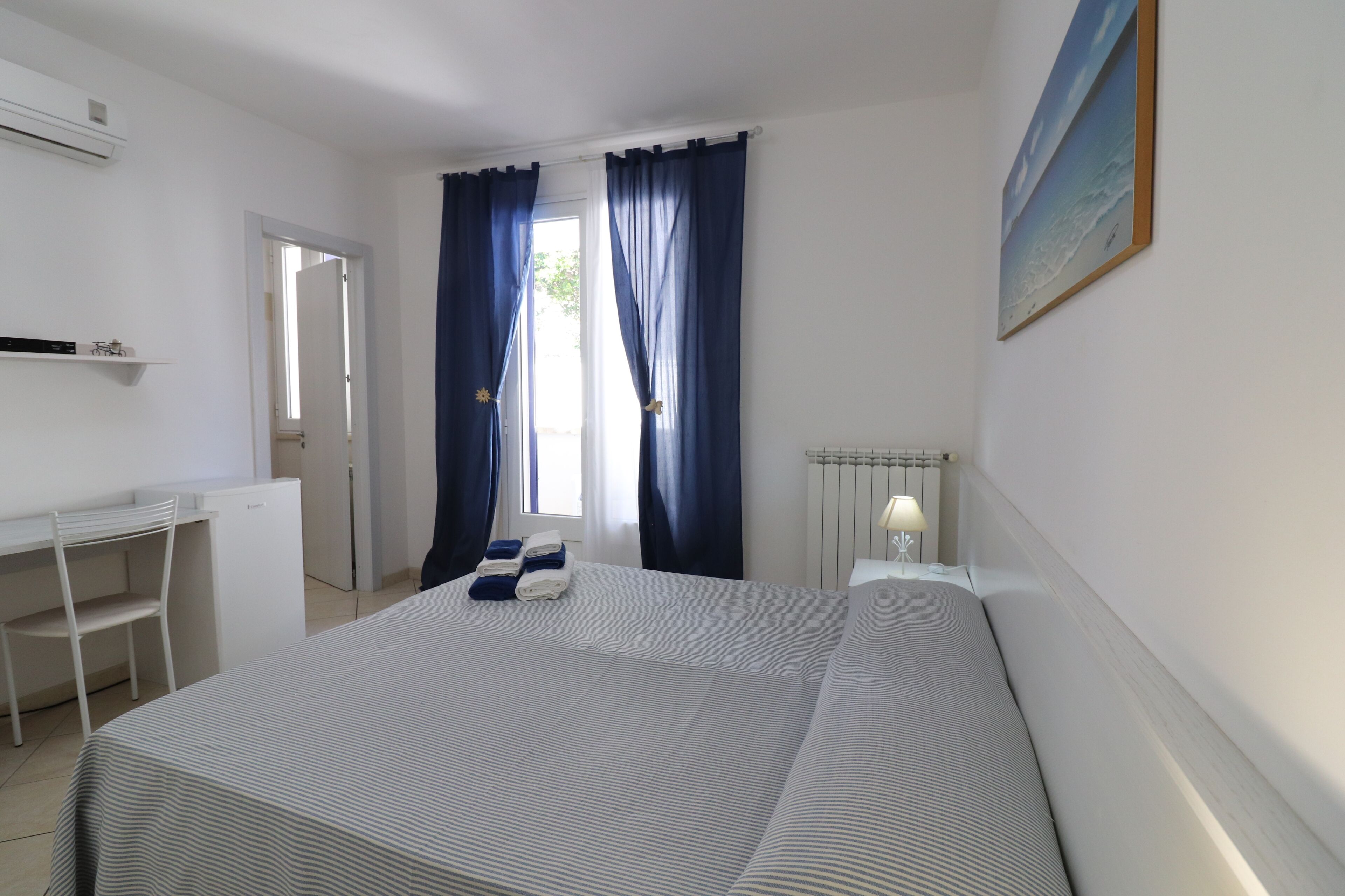 Un dormitorio con cama, escritorio y silla. Se ve una ventana con cortinas, un radiador y un cuadro en la pared.