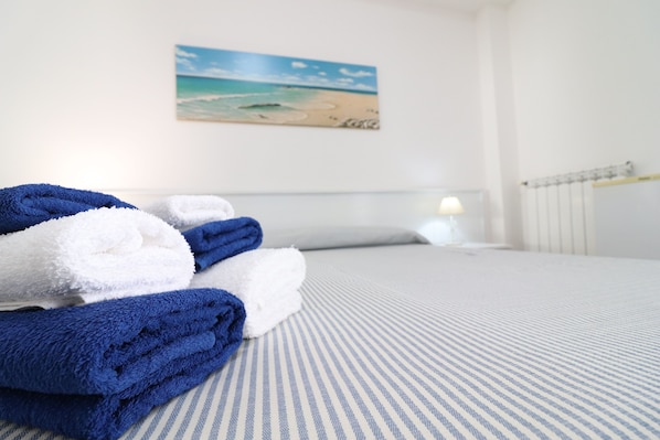Un lit bien fait, avec des serviettes pliées, un tableau représentant une plage accroché au mur et un radiateur.