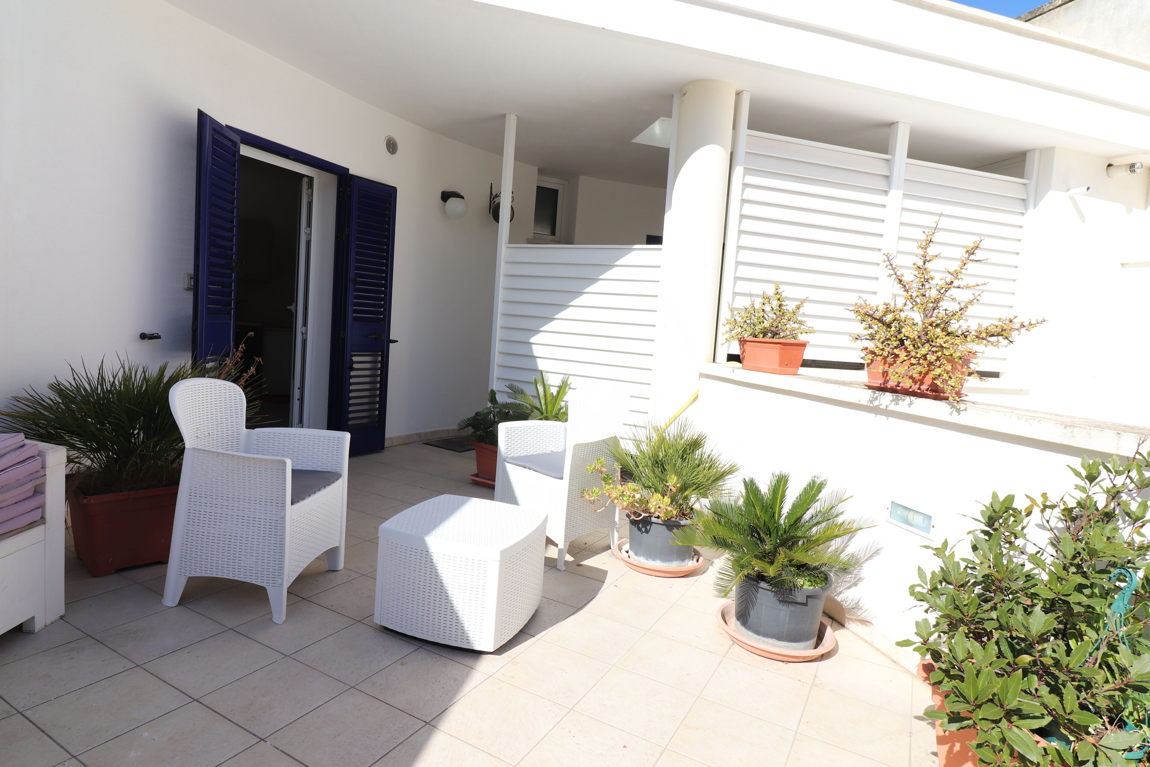 Un espace extérieur abrité, aménagé avec des meubles en osier blanc, des plantes en pot et un bâtiment blanc en arrière-plan.