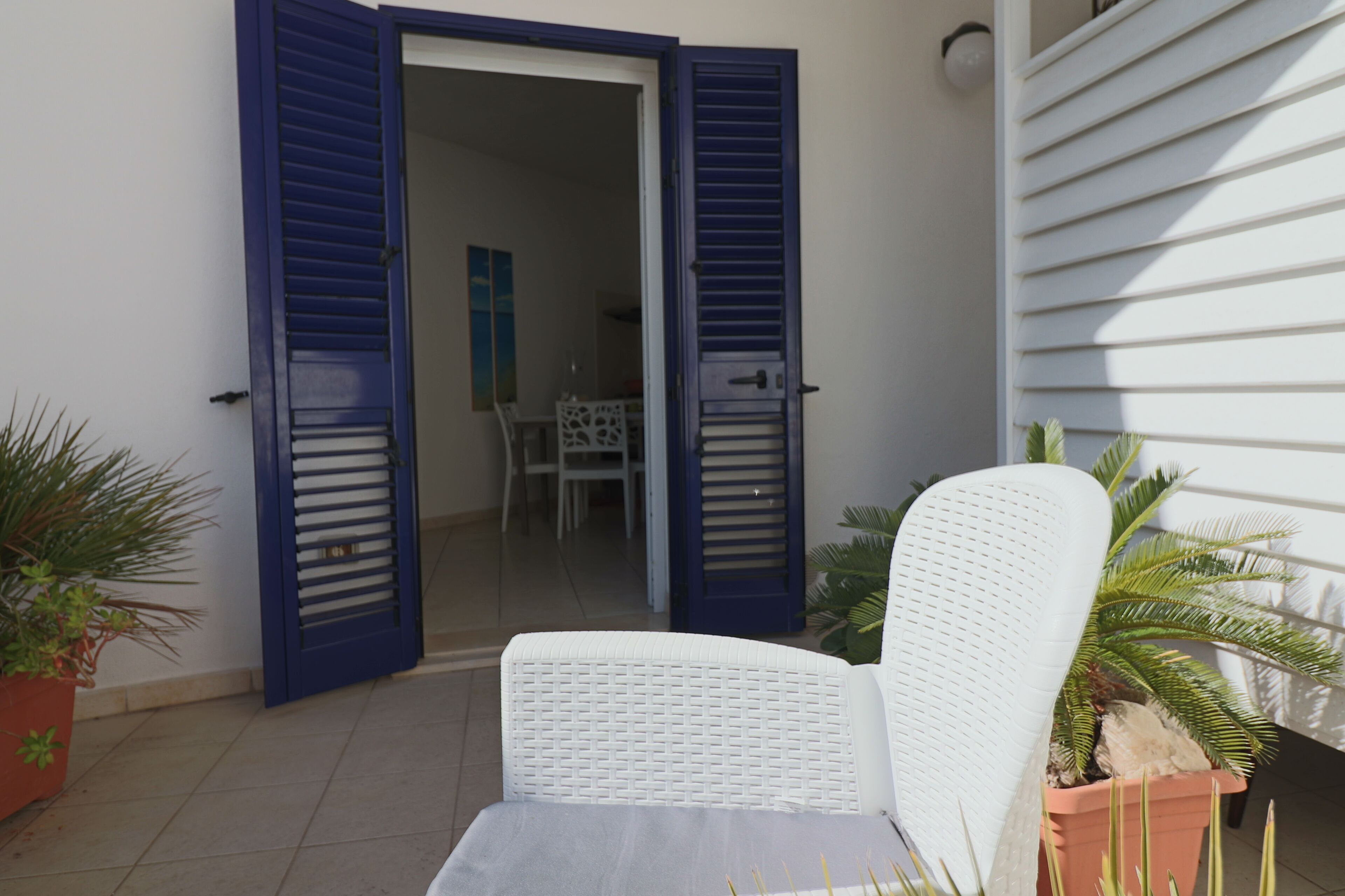 Puerta corredera de vidrio abierta que da a un balcón con una silla de mimbre blanca y plantas en macetas.