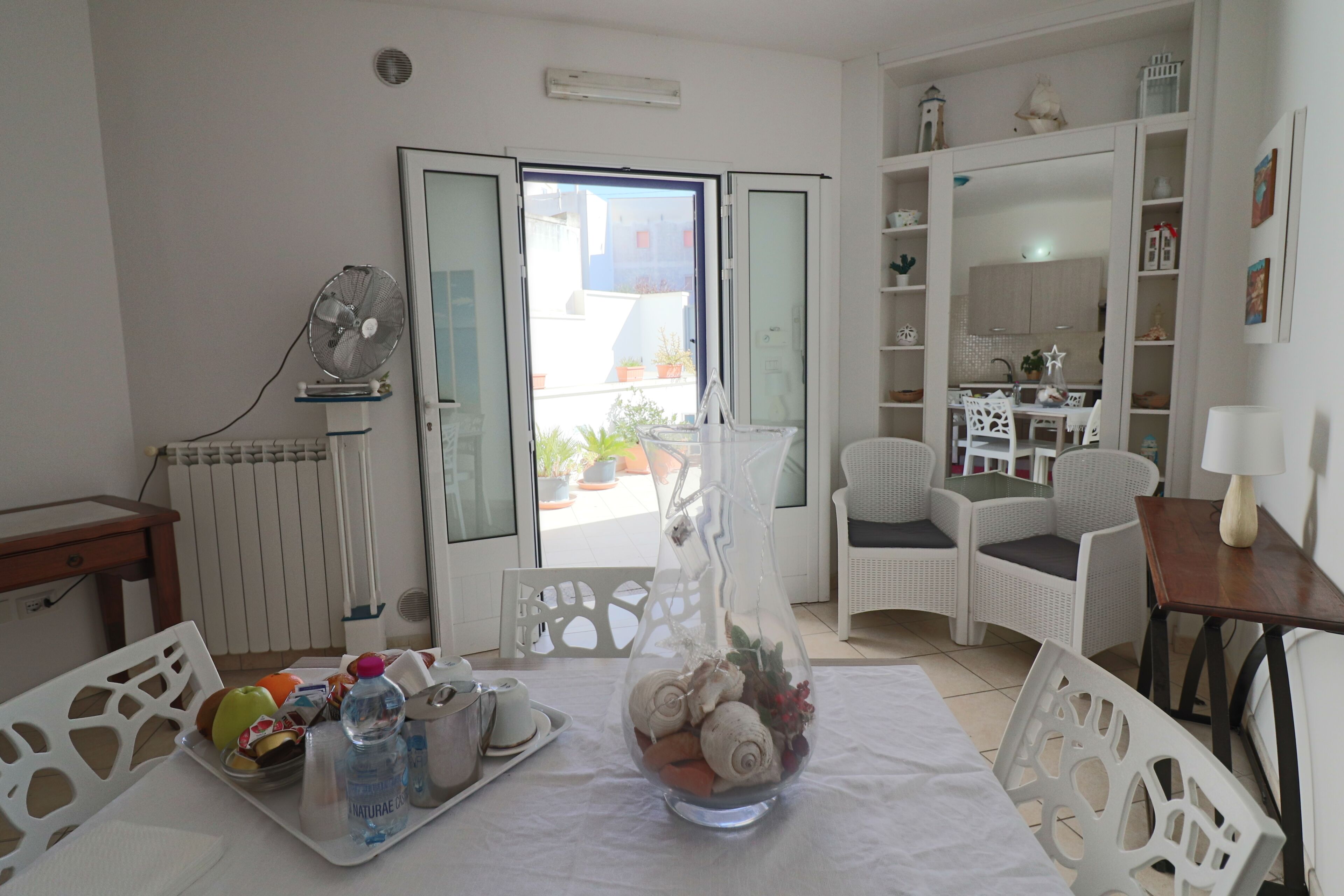 Camera d'albergo con tavolo apparecchiato per la colazione, una sedia in vimini, una poltrona bianca e vista sul balcone esterno.