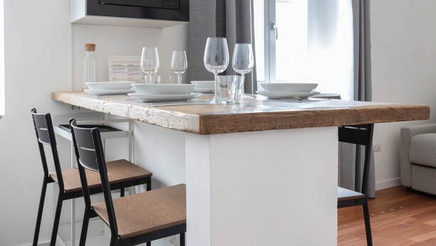 A kitchen island with a rustic countertop, white walls, and a window with curtains. There are three chairs, a bottle, and several wine glasses on the island. - Italianway - Umbria 50 B (Milan)