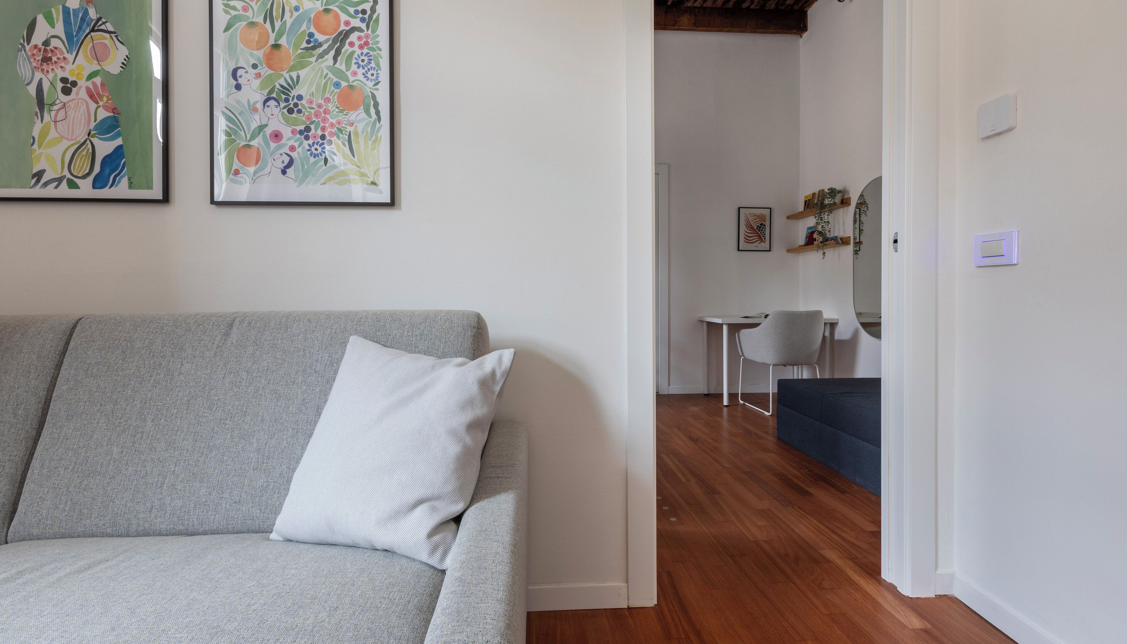 Una sala de estar con un sofá gris y un cojín blanco, una mesita con una silla, y una puerta que da a otra habitación.